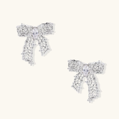 Elegant silver bow-shaped earrings with sparkling zirconia crystals and dangling ribbon details on cream background.
