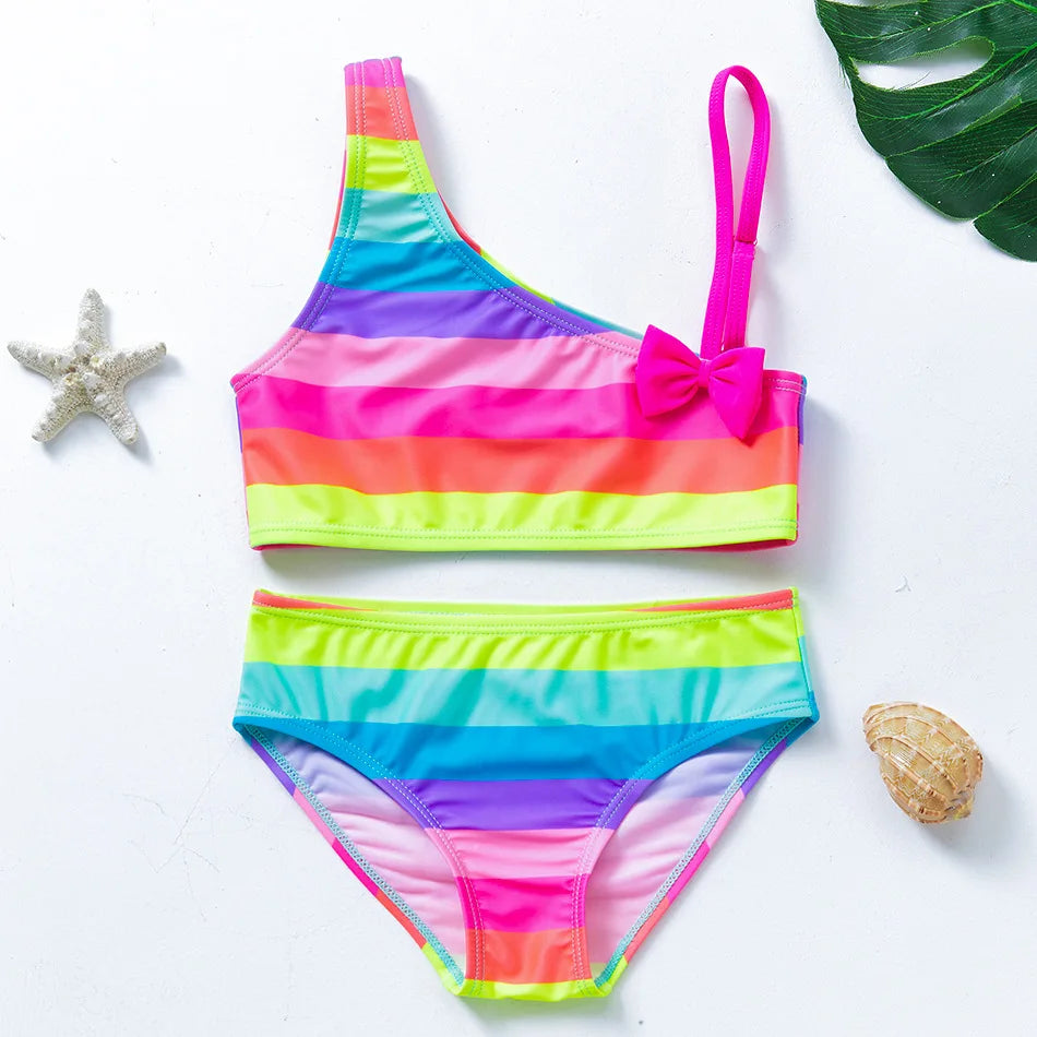 Two-piece rainbow striped girl's swimsuit featuring a golden crown 'Queen' graphic and a pink decorative bow.