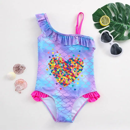 One-piece girl's swimsuit with purple and blue fish scale patterns, a colorful heart mosaic, and hot pink ruffles.