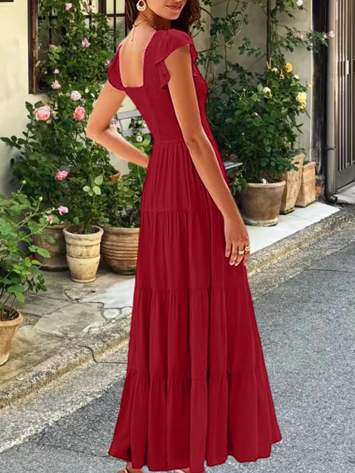 Woman from behind wearing a deep red tiered maxi dress with flutter sleeves and ruched waist in a garden setting.
