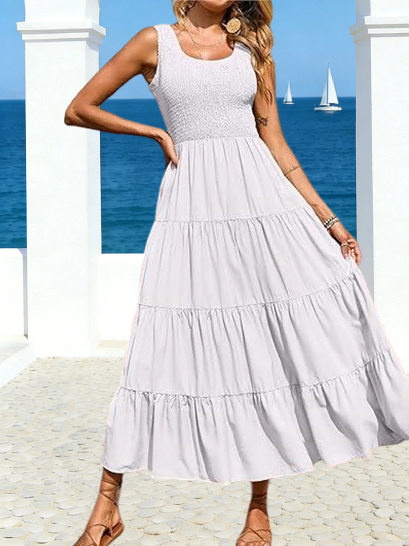 Woman wearing a white tiered smocked maxi dress standing on a sunny beach with sailboats in the background.